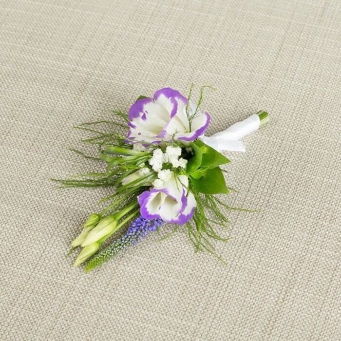 A refined wedding boutonniere is captured in close detail, resting on a pale beige linen surface whose soft, natural texture gives a calm, understated backdrop. The arrangement is centred on two open lisianthus flowers, their petals a pure, creamy white edged in a bold ring of velvety purple, each ruffled fold catching the light and giving the blooms a luxurious, almost hand-painted appearance. Around these focal flowers cluster small, bright white filler blossoms, similar to baby's breath, giving a gentle, airy fullness that softens the strong bi-colour contrast. Rising alongside them is a slim stem of muscari-like flowers, tightly stacked and bell-shaped, in a deep purple-blue shade that intensifies the palette and adds a vertical, textural line to the compact design. Threading in and out behind the flowers is a halo of fresh greenery, combining fine, feathery fronds reminiscent of asparagus fern with slightly broader leaves, so the foliage feels both structured and delicately wild, like something gathered from a shaded corner of a Catford garden near Ladywell Fields. Two pale green buds nestle at one edge, still closed and slightly curved, suggesting new beginnings and the quiet anticipation of the day. At the base, all the stems are firmly aligned and wrapped in a smooth band of crisp white satin ribbon, finished with a neat, narrow bow that signals professional craftsmanship and readiness to be pinned onto a lapel. The mood is elegant and composed yet gently romantic, ideal for a groom or groomsman at a local Catford ceremony who wants a subtle touch of colour and texture to complement a classic suit. This is the sort of detailed, hand-finished buttonhole a florist at Catford Flowers would prepare with care for a meaningful celebration.