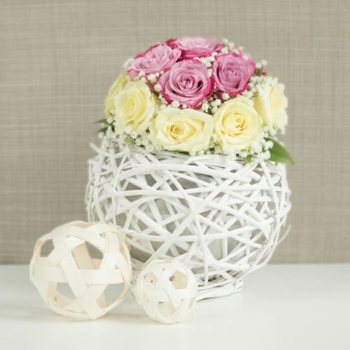 This image shows a graceful floral centerpiece arranged with the care and precision you might expect from an experienced Catford wedding florist preparing a table display for a local reception. At its heart is a carefully sculpted mound of roses, about a dozen in total, positioned so closely that their petals almost brush like silk. Half of the blooms are a soft, romantic pink with a hint of lavender, while the others are a warm cream that deepens toward buttery yellow at the centre, giving the whole piece a gentle, sunlit glow. The interplay of these two colours feels soothing rather than bold, evoking relaxed celebrations and intimate toasts rather than grand drama. Wisps of white baby's breath weave among the roses, their tiny clustered flowers lifting the design and bringing a feathery, cloud-like texture, similar to fine lace edging on a bridal gown. The flowers are arranged in a striking spherical vase, crafted from thin white-painted branches or wicker that twist and cross to create an intricate, open-work globe. Through its gaps you can glimpse the inner container, yet the overall impression remains light and sculptural. The vase rests on a spotless white tabletop, its simplicity amplifying the purity of the palette. On the left side of the frame, two decorative spheres made from pale, natural wood strips sit casually, one slightly larger than the other, echoing the woven structure of the vase but in a warmer tone. Behind, a neutral beige or light taupe wall with a faint fabric-like texture offers a calm, unobtrusive backdrop. The entire scene feels peaceful and warmly welcoming, perfect for a wedding breakfast near Catford Bridge station or as a sophisticated centrepoint for a family gathering at home.