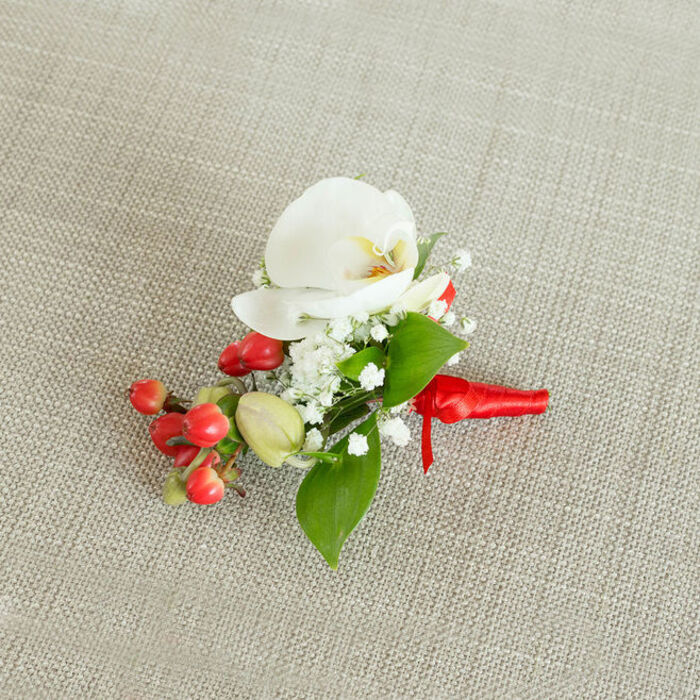 This image shows a single, beautifully constructed boutonniere or small corsage set against a subtly textured, light beige woven fabric, similar to natural linen you might see in a Catford artisan market stall. The composition is captured from a slightly elevated angle, allowing the entire floral piece to be seen in clear detail. At the centre is a pristine white Phalaenopsis orchid, its petals broad, velvety, and gently curving outward, creating a serene, butterfly-like silhouette. Nestled within its throat is a soft blend of creamy yellow and light orange, offering a warm focal point that contrasts delicately with the cool white of the petals. Encircling the orchid, abundant clusters of white baby's breath form a soft, cloud-like halo, each tiny floret contributing to a sense of airiness and romance. Interspersed through this cloud are smooth green leaves in different shapes and sizes, from broader, pointed blades to more slender, elongated forms, introducing a fresh, botanical depth that feels as natural as a stroll through Forster Memorial Park. On one side of the arrangement, a compact spray of bright, glossy cherry-red berries, most likely hypericum, adds a lively pop of colour, their rounded forms and polished surfaces suggesting plumpness and vitality. Nearby, a few paler green buds echo the same shape in a more understated tone, hinting at developing fruit and gentle seasonal change. The stems are carefully aligned and bound with a rich, red satin ribbon, wound tightly and tied into a neat, understated knot with short ends resting smoothly against the woven backdrop. The soft, even daylight highlights every surface-from the orchid's satin sheen to the subtle weave of the fabric-conveying a sense of quiet, considered elegance ideal for weddings, formal dinners, or special celebrations in and around Catford.