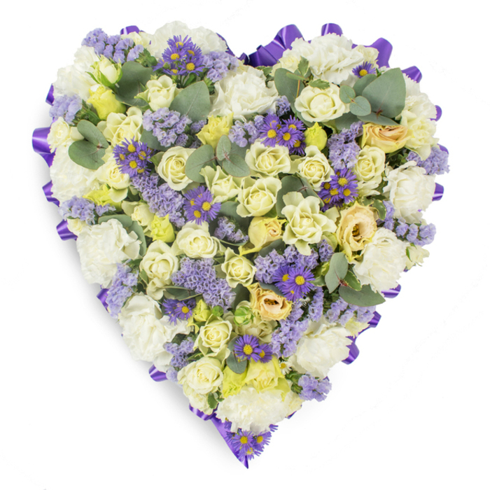 Captured on a pure white studio surface, this heart-shaped sympathy arrangement reads as a quiet, elegant ode to memory. The design is densely filled with soft white carnations and a chorus of cream to pale yellow roses, their petals layered from tight centers to gently unfurling edges, building a plush, tactile landscape that invites a tender touch. Interspersed are clusters of delicate lavender statice and small purple daisy-like flowers with notable yellow centers, which create shimmering threads of cooler color and add visual rhythm across the pale field. Scattered throughout are smooth, rounded eucalyptus leaves in blue-green tones; their matte sheen and distinctive silhouette provide subtle structural pauses among the blooms and suggest a faint, refreshing scent that complements the floral notes. The heart is neatly bordered by a continuous, deep purple satin ribbon, folded into crisp loops that define the silhouette and add a formal, respectful finish to the tribute. Soft, diffused lighting emphasizes the gentle tonal transitions and the layered textures - velvety rose petals, frilled carnations, and papery statice - while the overall feeling remains composed and consoling. Intended for use at wakes, memorial services, or as a condolence gift placed on a family altar, this arrangement would suit services across Catford and nearby precincts such as Ladywell or Brownhill Road, offering a quiet symbol of devotion and heartfelt sympathy during a time of loss.