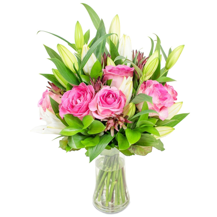A meticulously arranged bouquet stands in a simple clear glass vase against a white background, its composition feeling both abundant and precisely balanced. Dominant in the centre are several large pink roses, their petals layered in sumptuous waves of blush and deep fuchsia that create a tactile, velvety surface; the roses read as romantic and full-bodied. Interspersed among them, white lily buds sit tall and elegant - many still closed, some beginning to open into trumpet-like forms with faint green tips - promising more bloom and a soft, airy scent. Surrounding this core is a rich mix of foliage: broad pointed leaves add weight and a glossy contrast, while finer, lighter green sprigs fill gaps and introduce gentle movement. Small, darker reddish-brown filler elements pepper the bouquet, lending depth and a whisper of rustic character against the otherwise fresh palette. The clear vase, slightly tapered and holding transparent water, exposes the trimmed stems below and affirms the arrangement's freshness and care. Soft overhead lighting highlights petal edges and the sheen of the leaves, producing a clean, catalogue-ready image that still feels warm and hand-crafted. This bouquet would make a considered gift for anniversaries or birthdays and is suited to brightening a living room or reception near Catford Bridge or a kitchen table by Mountsfield Park - a floral statement that combines everyday elegance with the attentive touch of a local florist.