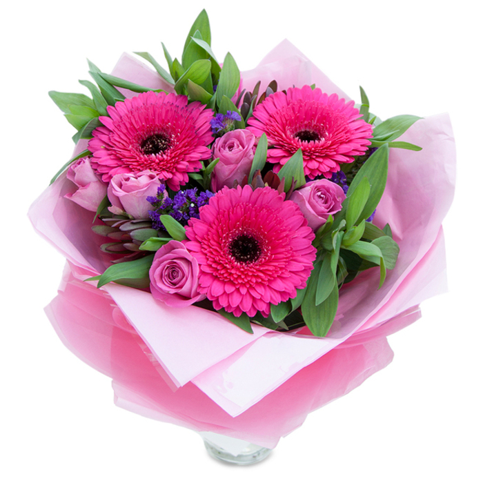 I love how this bouquet balances playfulness with quiet refinement: three vivid gerberas in fuchsia act like joyful exclamation marks, their many petals radiating from dark centres that anchor the colour story. Complementing them are roses in a muted lavender-pink or dusty rose shade, each bloom presenting layered, velvety petals that open with a tender, sculptural grace. Interspersed throughout are small sprays of purple statice, their tiny clustered blossoms lending a whisper of wildflower charm and textural contrast against the smoother petals. A generous weave of greenery-broad salal-like leaves and hints of eucalyptus-frames the flowers with a crisp, slightly dewy character that speaks of freshness and careful sourcing. The bouquet is wrapped with an inner cushion of soft pale pink tissue and an outer veil of sheer fuchsia-pink cellophane that shimmers subtly, the wrapping folded with the precision of a neighbourhood florist preparing a keepsake delivery. The visible rim of a clear glass vase at the bottom signals that the arrangement is meant to be displayed immediately, filling a room with colour and a faint bouquet-scent of roses and green stems. Picture this on a coffee table in a flat near Mountsfield Park or brightening a kitchen window on Catford Broadway-an intimate, celebratory piece ideal for anniversaries, heartfelt thank-yous, or simply to make someone's day a little lighter.