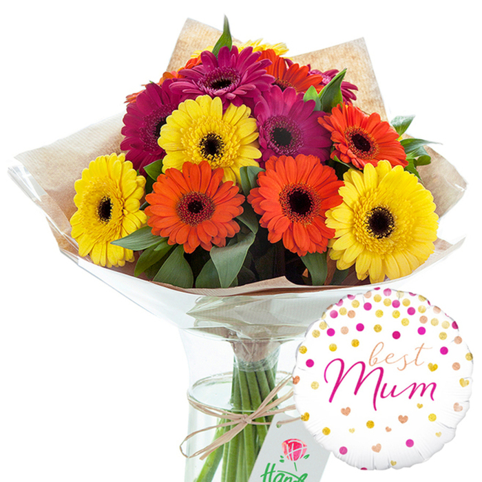 In this carefully composed studio image, a generous bouquet of gerbera daisies stands tall in a clear glass vase, the whole scene set against a plain white background so every colour and texture can be appreciated. About a dozen large gerbera blooms form a rounded, hand-tied arrangement, each flower perfectly open with its signature daisy shape. The petals create bold rings of colour in three main tones: sunny, butter-yellow, lively orange with a soft amber warmth, and intense magenta that leans into deep fuchsia, reminiscent of vivid shopfront displays along Rushey Green. Each bloom has a distinct dark central disk, adding definition and drawing the eye inward before the bright petals carry it back out again. Rich green foliage is woven carefully among the flower heads, the leaves adding both volume and an earthy contrast to the more playful colours. The stems are gathered and wrapped in a layer of rustic, slightly crumpled brown kraft paper that has a tactile, natural look, like something you might carry home from a Catford market stall. Over this sits an outer layer of clear, glossy cellophane that hugs the form of the bouquet and reflects subtle highlights from the studio lighting. Around the stems, a piece of natural twine is tied in a practical yet charming bow, emphasising the hand-crafted feel of the design. The transparent glass vase reveals straight, fresh green stems standing in clear water, confirming the bouquet's freshness. To the lower right, a round white foil balloon adds a distinctly celebratory feature. Its surface is sprinkled with metallic gold and pink dots and tiny hearts, suggesting scattered confetti at a family gathering. In the centre, in flowing pink script, appears the affectionate dedication best Mum, turning this already vibrant floral arrangement into a complete gift that expresses gratitude, affection, and warm celebration for a mother or cherished mother figure in Catford.