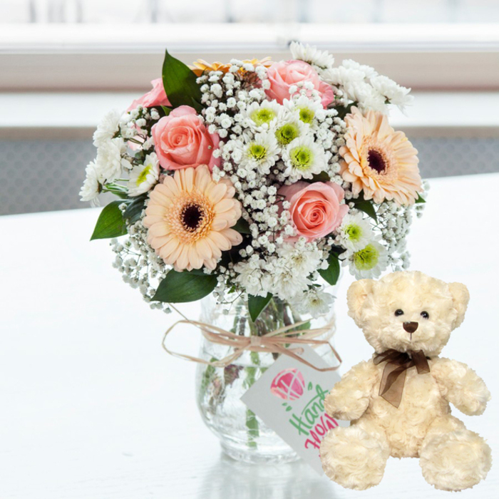 A soft, romantic still life unfolds on a pristine white table, reminiscent of a quiet corner in a Catford home just off Rushey Green. In the centre stands a clear glass jar vase, simple and rustic, filled with a fresh pastel bouquet that seems to glow in the natural daylight. Inside the vase, stems of varying thickness create a tapestry of greens, carefully arranged so the blooms above fan out in a rounded, generous shape. Dominating the arrangement are pale pink roses, their petals opening in gentle spirals, and peach-toned gerbera daisies whose large, flat faces are accented by deep brown centres that add a subtle, earthy contrast. Nestled among them, airy sprigs of white baby's breath float like tiny stars, softening the lines between each flower. White chrysanthemums, each with a neat halo of petals around a lime-green centre, frame the bouquet, lending it a clean, uplifting brightness. Around the neck of the vase is a wrap of natural twine, tied casually yet deliberately into a bow. From this twine hangs a petite, rectangular white card featuring a simple pink heart motif and delicate green handwriting, suggesting a handmade, heartfelt sentiment-perhaps an anniversary note or a thank-you message. To the right, a cream-coloured teddy bear sits upright on the same white surface. Its plush fur appears fluffy and touchable, with small dark eyes, a modest brown nose, and a narrow, dark brown satin ribbon tied neatly at the throat. The background is softly out of focus, flooded with gentle light, so the viewer's gaze lingers on the pairing of flowers and bear. Together they create a tender tableau of affection and comfort, perfect for celebrating love, sending get-well wishes, or surprising someone in Catford with a same-day delivery that feels deeply personal.