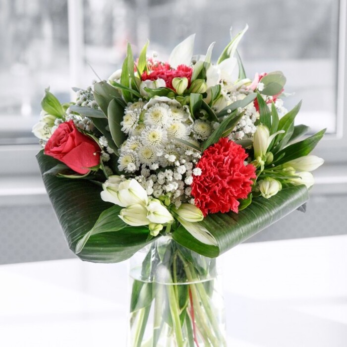 A beautifully composed red and white bouquet sits in a clear cylindrical glass vase, the kind a local Catford florist would place at the centre of a dining table for a special family gathering. The vase is filled with clean water, making the straight, neatly cut green stems clearly visible and giving a reassuring sense of freshness and professional care. Above the glass, the flowers gather into a rounded, generous dome of colour and texture. The most striking elements are the deep red roses, velvety and full, their gently unfurling petals echoing the look of classic romance and heartfelt devotion. At the front, a large, deeply ruffled crimson carnation stands out; its tightly layered frills catch the light and create a rich, textured focal point. Between and around these red blooms, a selection of white and creamy flowers offers calm contrast. White alstroemeria with fine speckles at their throats, daisy-like white blossoms, and the presence of several unopened white lily buds together bring an elegant softness and the promise of future fragrance as the days go by. Delicate sprays of white gypsophila weave through the bouquet, their tiny star-shaped flowers floating like a fine mist, adding airiness and a slightly dreamy quality. The arrangement is framed and supported by an abundance of greenery: broad, dark green tropical leaves are carefully curled and wrapped around the base, forming a smart, structured collar, while lighter, eucalyptus-like sprigs and tapering green leaves create layers of texture and fresh colour. The whole bouquet rests on a pristine white surface against a softly blurred, light background, probably a bright wall or window, so the flowers themselves command all attention. It feels perfect for marking meaningful moments in Catford-an anniversary dinner, a birthday near Ladywell Fields, or a thoughtful delivery to brighten a home office-radiating joy, appreciation, and warm affection in every petal.