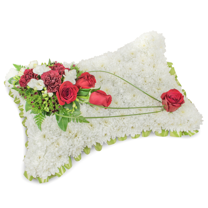 Presented against a clear, white backdrop, this rectangular floral pillow is a classic expression of sympathy and remembrance, its entire surface formed from hundreds of compact, pristine white chrysanthemum florets that create a soft, cloudlike texture. The densely arranged heads produce a subtle, domed silhouette and a tactile surface that reads as both pure and soothing. A thin border of pale green ribbon and glimpses of foliage run around the perimeter, quietly finishing the shape with a hint of fresh colour. Anchoring the composition on the upper left is an elaborate cluster of deep red blooms - full, velvet-petalled roses in different stages of opening paired with richly hued crimson carnations whose layered, ruffled petals bring shadow and rhythm. Little sprays of delicate white blooms, possibly freesias or small spray chrysanthemums, sit among the reds to add contrast and a whisper of light. A background of lush green leaves and fine fern fronds supports the main cluster, and three elegant strands of bright green bear-grass sweep away from this focal point toward the lower right, drawing the eye along a gentle diagonal. The sweep concludes at a single, perfectly shaped red rose placed near the bottom right corner, a poignant focal note suggesting enduring affection. The soft studio light emphasises the velvety reds against the pure white cushion, and the overall mood is one of measured dignity and solace. This arrangement would be fitting for a funeral or memorial in Catford Bridge or at a local church near Rushey Green, offering a respectful and comforting floral tribute.