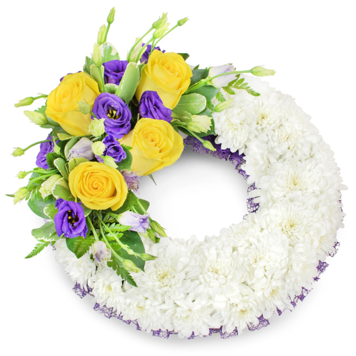 A round memorial wreath presented on a stark white surface, its design grounded in classic sympathy floristry and careful textural balance. The principal structure is made up of a thick band of pristine white spray chrysanthemums-tiny, multi-petalled blooms packed so closely they form a continuous, pillowy ring that diffuses light and suggests peacefulness. From the upper-left side springs a thoughtful cascade of colour: vivid yellow roses with petals curled back in soft spirals, paired with rich purple lisianthus whose velvety petals form graceful cups. A scattering of unopened lisianthus buds in soft lavender and pale green punctuates the spray, giving the eye places to rest and implying quiet continuity. Mixed greenery-glossy oval leaves, lightly variegated foliage, and delicate fern fronds-frames the coloured cluster and enhances the brightness of the blooms. Along the wreath's inner and outer curves a slender, dark purple mesh trim peers out from beneath the chrysanthemums, mirroring the deep lisianthus and lending a subtle handcrafted finish. The overall mood is poised and consoling: clean white for purity and memory, yellow for gentle warmth, purple for solemn beauty. As a local floral designer serving Catford and the surrounding Lewisham neighbourhoods, I imagine this wreath arriving at a chapel near Catford Bridge or being placed with quiet dignity at a service close to Mountsfield Park, conveying respect, comfort and heartfelt farewell.