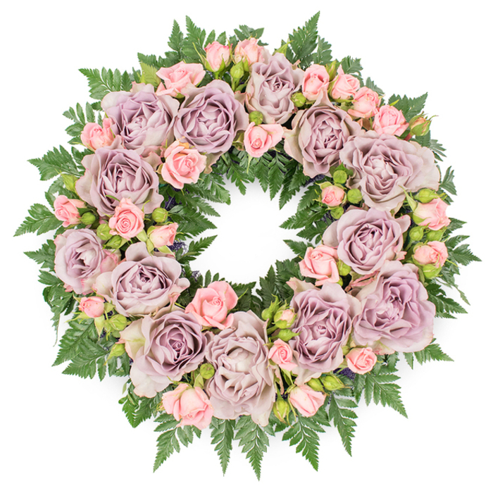 Viewed directly from above against a bright white background, this circular funeral wreath reveals an exquisite balance of colour, texture, and meaning. The design is densely ringed with large, fully open roses in a muted spectrum of dusty lavender, slate-tinged mauve, and soft greyish-purple, each blossom unfolding in an intricate maze of petals that spiral inward toward a gently shadowed core. Between these stately blooms sit groupings of delicate blush pink spray roses, some in full flower, others only half open, and a scattering of tiny buds just beginning to part. This variety gives the wreath a dynamic, almost breathing quality, like a collection of moments across a lifetime. Beneath and around the roses, a thick bed of dark green fern fronds fans outward, the serrated edges and fine segments of the leaves forming a rich, verdant border. This greenery peeks through the flowers in small arcs and hints, grounding the pastel palette with a strong sense of nature and continuity. Soft, neutral lighting falls evenly across the wreath, bringing out the subtle gradations in purple and pink while preserving a calm, contemplative mood. The overall impression is one of refined elegance and quiet sympathy, the type of tribute often chosen for funerals and celebrations of life held near Catford's churches or local crematoria. It feels like the work of Catford Flowers, or another skilled local florist, who knows how to arrange blooms so that grieving families see not just flowers, but a circle of remembrance that reflects dignity, peace, and enduring affection.