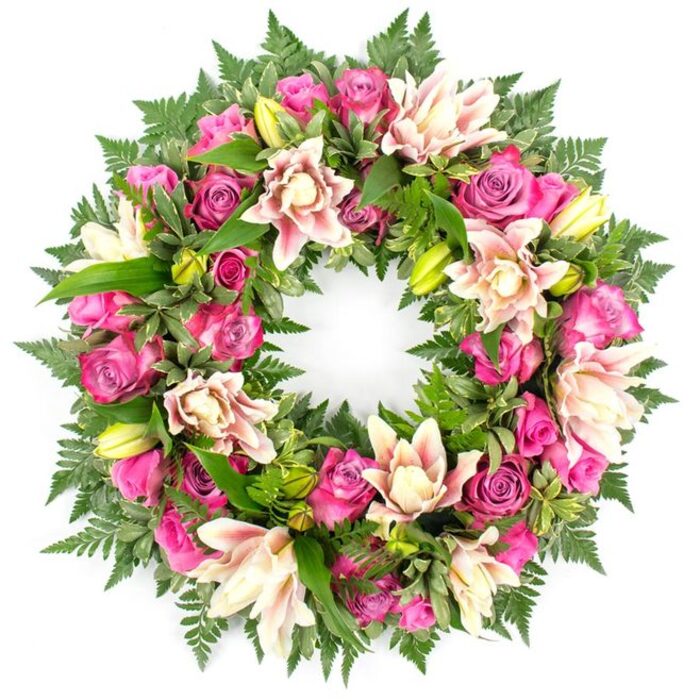 Displayed on a bright white background, this circular funeral wreath looks both opulent and serene, meticulously arranged so every flower and leaf feels intentionally placed. The ring is completely covered with blooms, with no bare spaces, creating a full, luxurious appearance that draws the eye around the entire circle. Dominating the design are roses in rich berry tones, from deep magenta to lighter fuchsia and tender pastel pink. Their petals are plush and velvety, spiralling tightly at the centre of some blooms while forming open rosettes in others, capturing the natural variation you see in fresh flowers just delivered that morning. Interwoven between these roses are large double lilies, their layered petals opening wide in shades of creamy white, soft blush, and hints of pale peach, some petals touched with a slightly deeper pink hue near the centre. A few upright, lime-green buds sit among them, their pointed tips giving a subtle vertical accent amid the rounded heads of the roses. The flowers are anchored in a thick base of foliage: dark, feathery fern fronds overlap like delicate lace, while broader, glossy leaves nestle beneath and behind the blooms, adding both depth and contrast. The rich greens intensify the warmth of the pinks and creams, making the colours appear even more vibrant against the stark white background. This wreath feels ideal for a farewell service in Catford, perhaps placed by the coffin or at the front of a local chapel, offering a respectful, elegant expression of sympathy that wraps grief, gratitude, and memory into one continuous floral embrace.