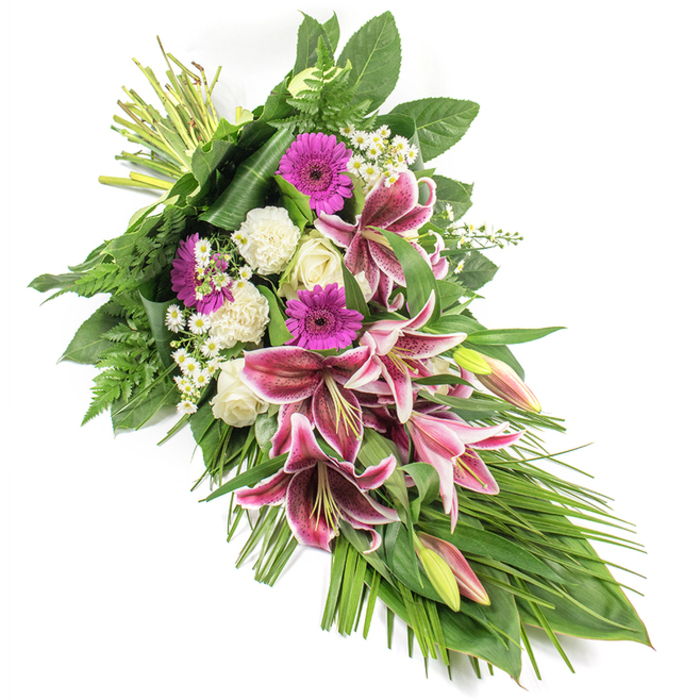 A refined funeral spray, styled as an elongated teardrop sheaf, is shown against a crisp white background, its colours and textures standing out with calm intensity. The arrangement runs diagonally from top left to bottom right, as if gently resting on an invisible casket or memorial table, a style often requested by families in Catford for farewell services. At its heart are several commanding Stargazer lilies, their petals a vivid fuchsia-pink with precise white margins and deep burgundy speckling, some flowers fully unfurled, others still partially closed, suggesting a life remembered through many stages. Nestled between them are bold magenta gerbera daisies, their round faces offering a modern contrast and a note of warmth. Clusters of white roses, many in graceful bud form, bring a classic, timeless elegance, while soft, ruffled white carnations infuse the design with a feeling of tenderness and affection. Small daisy-like white filler blooms are scattered throughout, like tiny points of light, softening the transitions between the larger flowers. The entire spray is framed by lush greenery: broad, dark leaves provide a polished structure; fronds of fern add feathery texture; and long, slender blades of foliage sweep outward and downward, accentuating the cascading form. The bound stems at the top left reveal the hand-tied craftsmanship, as though freshly prepared by a Catford florist ready to deliver to a nearby chapel or home. The overall mood is one of quiet strength, gentle colour, and respectful beauty, making this arrangement a fitting tribute to express sympathy, love, and remembrance during moments of farewell.