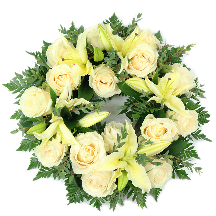 This carefully crafted circular wreath, photographed flat against white and viewed from above, pairs creamy white roses with soft pale yellow lilies to form a composed, reverent memorial piece. Approximately fifteen roses, fully open with their classic inward spirals, create a serene ivory canvas; interspersed are several trumpet-shaped lilies-some opened to reveal their pale yellow throats and graceful stamens, others held as refined buds-adding vertical interest and a hush of expectancy. The blooms are set on a generous base of greenery: airy fern fronds lend a feathery border, while broader, rounded leaves-suggestive of eucalyptus-introduce cooler, slate-green tones that deepen the design's dimension. The overall composition is symmetrical and dense, the flowers arranged close together so the surface appears plush and continuous, the circular outline deliberate and unbroken to symbolise continuity, memory, and the cycle of life. Lighting is soft and even, allowing the cream and lemon hues to glow subtly against the darker foliage and emphasizing the wreath's tranquil character. Texturally the piece balances smooth rose petals, the satiny curves of lily sails, and the lacy texture of fern, inviting a contemplative, almost tactile response. Made with the sensibility of a neighbourhood florist serving Catford and nearby Mountsfield Park, this wreath conveys calm, purity, and respectful condolence-suitable for a funeral chapel, graveside setting, or as a sincere remembrance sent to comfort grieving families.