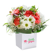 There's a gentle, uplifting energy that radiates from this Catford-inspired bouquet, elegantly nestled in a smart white square gift bag. A cascade of coral and rich red carnations, their plush, frilled petals brimming with subtle scent, gather alongside bright daisies-each white blossom crowned by a fresh lime-green center. The arrangement feels like a burst of optimism, reminiscent of the community spirit seen down Winslade Way, coming together in perfect harmony. Wispy trails of baby's breath weave between the blooms, lending a soft innocence that is further celebrated by a few discreet, purple-tinged buds quietly gracing one side-a nod to both tradition and the excitement of new beginnings. Framing the florals, lush deep green foliage provides a glossy, supporting embrace, sculpting the bouquet into a rounded display that seems to bloom with heartfelt intention. The sturdy ribbon handles and pure white of the bag hint at a prepared-for-anything gift, ready to be shared with loved ones across Sandhurst Road or the Catford Bridge area. The overall composition, handsomely lit on a pure white background, accentuates the bouquet's freshness, artistry, and local charm. Every stem and petal whispers a story of care-a wonderful choice for birthdays, heartfelt surprises, or simply brightening a quiet afternoon in SE6.
