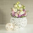 A vision of fresh beginnings unfolds in a Catford kitchen or lounge, with a floral arrangement that effortlessly blends handcrafted charm and gentle, springtime hues. Central is an airy white vase, ingeniously constructed from openwork rattan or light, painted branches, its spherical form making an elegant, almost floating base for the flourishing bouquet. Eustoma blossoms in soft cream and subtle yellow rise at the core, their ruffled petals beautifully contrasted by wisps of pink stock. At the design's base, lively purple fillers-perhaps aster or petite allium-emerge, lending texture and cheerful vibrancy. A ring of verdant green leaves encircles the florals, inspired by the leafy avenues stretching along Bromley Road. To one side, two decorative orbs, expertly woven from pale rattan, recall the artisanal spirit found at Catford's weekend markets. The arrangement sits atop a gleaming white surface, with the gentle, diffused morning light enhancing the bouquet's soft tones and natural forms. Behind, a delicate beige background with subtle fabric texture echoes the area's preference for understated, homely elegance. Calming and celebratory in equal measure, this composition by Catford Flowers feels perfect for marking a wedding breakfast, hosting friends, or bringing South London's creative warmth indoors, whatever the season.
